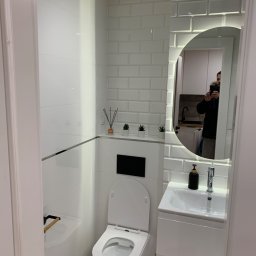 Efekthome - Nowoczesna, biała łazienka z toaletą wiszącą, umywalką w szafce i lustrem z podświetleniem LED. Ściany wyłożone białymi, prostokątnymi płytkami imitującymi cegłę. W tle odbicie osoby robiącej zdjęcie.