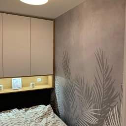 Efekthome - Sypialnia z szarą ścianą ozdobioną motywem liści palmowych, zabudową meblową nad łóżkiem z półką oświetloną ciepłym światłem, na której stoją dekoracje i ramka ze zdjęciem. Na suficie okrągła lampa.