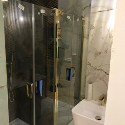 Efekthome - Nowoczesna kabina prysznicowa z złotymi profilami, zamontowana w łazience z marmurowymi i geometrycznymi płytkami, widoczna umywalka wolnostojąca i bateria.