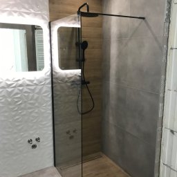 Efekthome - Wnętrze łazienki w trakcie remontu, widoczna kabina prysznicowa ze szklaną ścianką, szare płytki na ścianie i drewnopodobne na podłodze, białe trójwymiarowe płytki na ścianie obok, oświetlone...