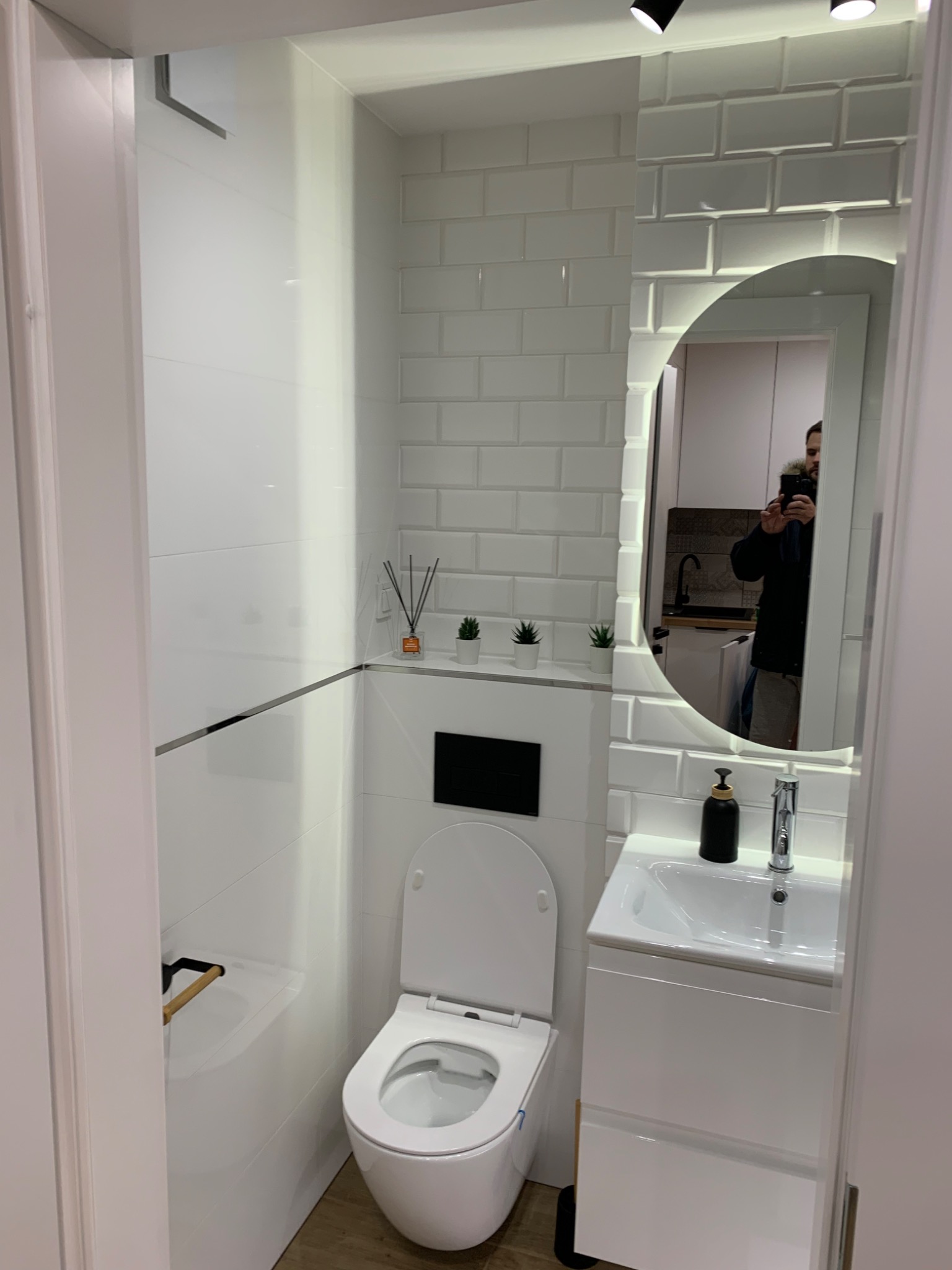 Nowoczesna, biała łazienka z toaletą wiszącą, umywalką w szafce i lustrem z podświetleniem LED. Ściany wyłożone białymi, prostokątnymi płytkami imitującymi cegłę. W tle odbicie osoby robiącej zdjęcie.