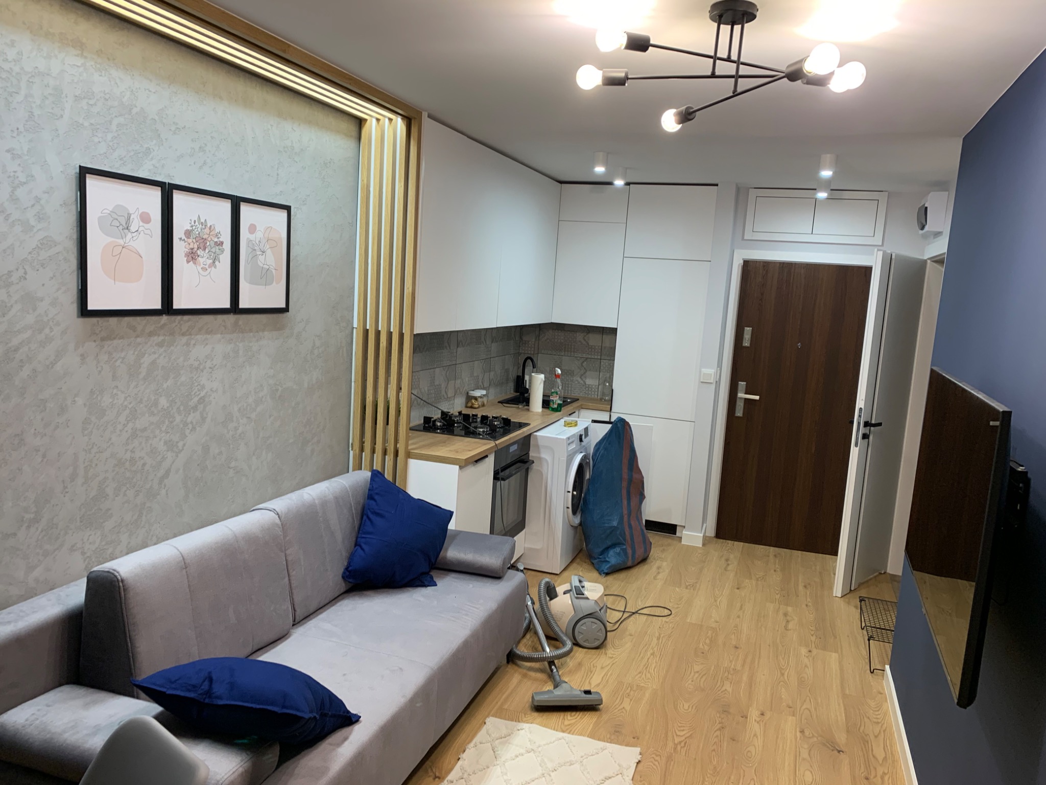 Wnętrze mieszkania po remoncie: szara ściana z trzema obrazami w czarnych ramach, szara sofa z niebieskimi poduszkami, białe meble kuchenne, drewniana podłoga, odkurzacz i pralka.