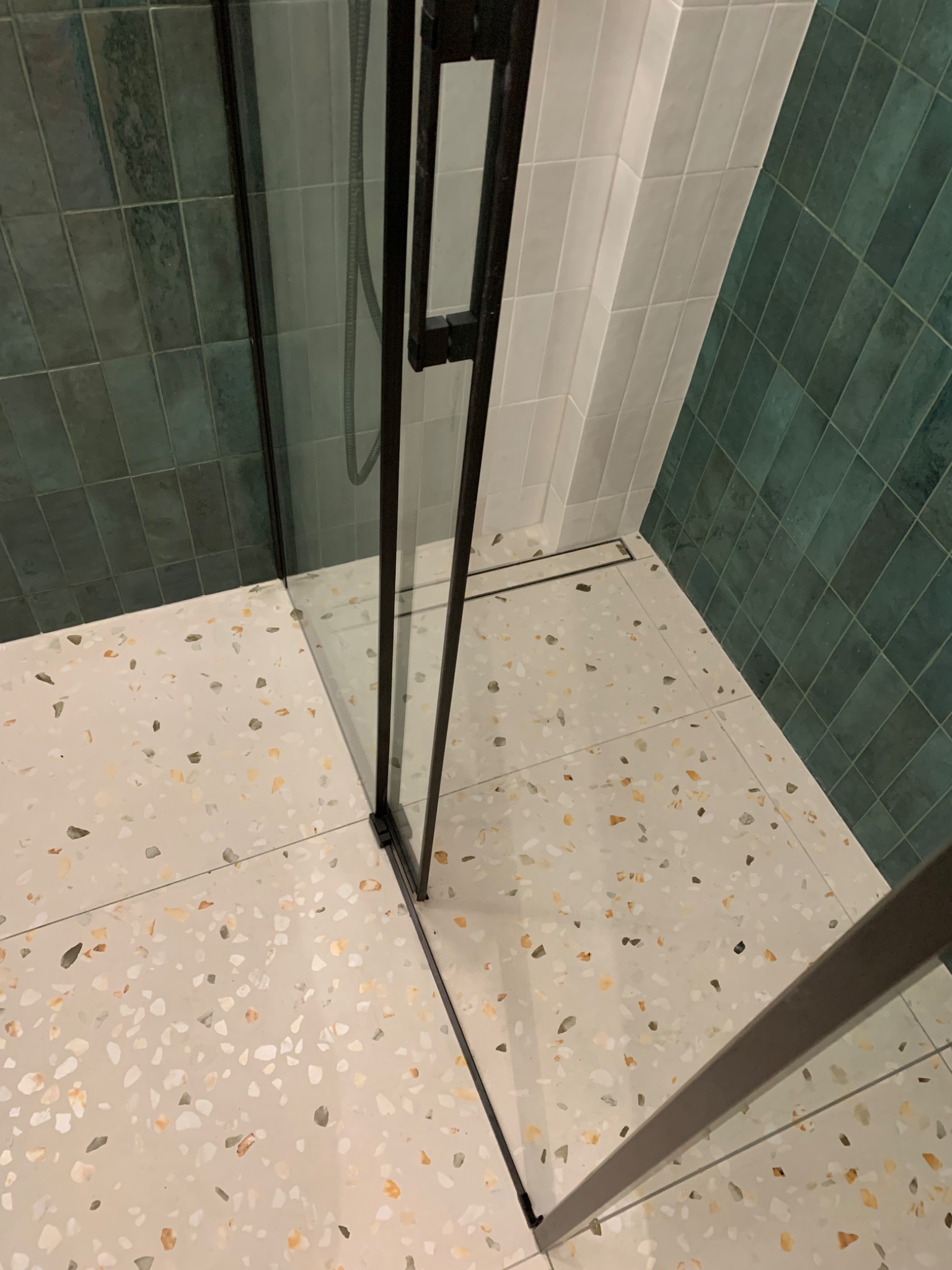 Nowoczesna kabina prysznicowa z czarnymi profilami, zielone i białe płytki na ścianach, podłoga w stylu lastryko, widoczny odpływ liniowy.