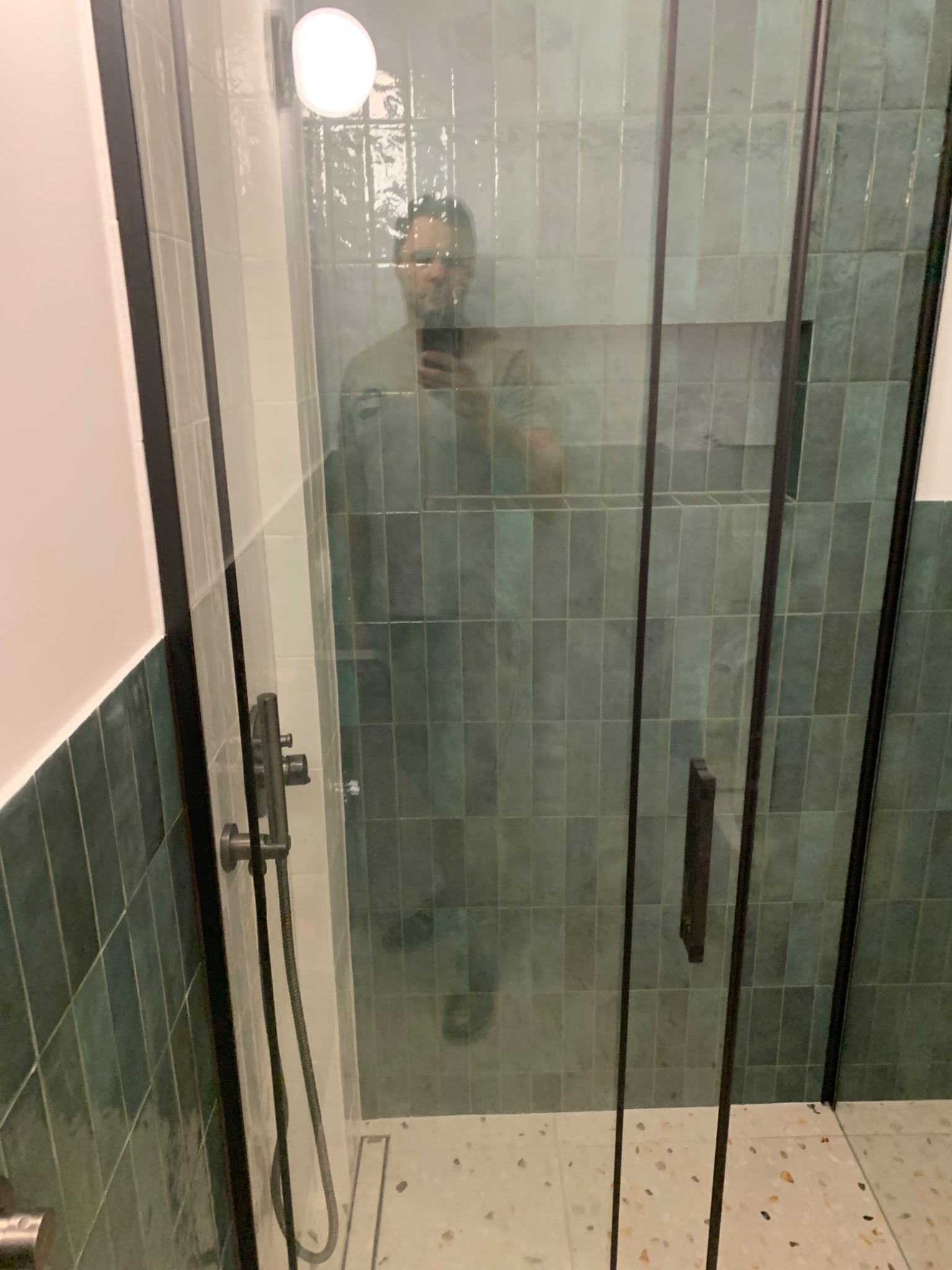 Nowoczesna kabina prysznicowa z ciemną ramą i zielonymi, pionowo ułożonymi płytkami, odbicie fotografa w szkle.