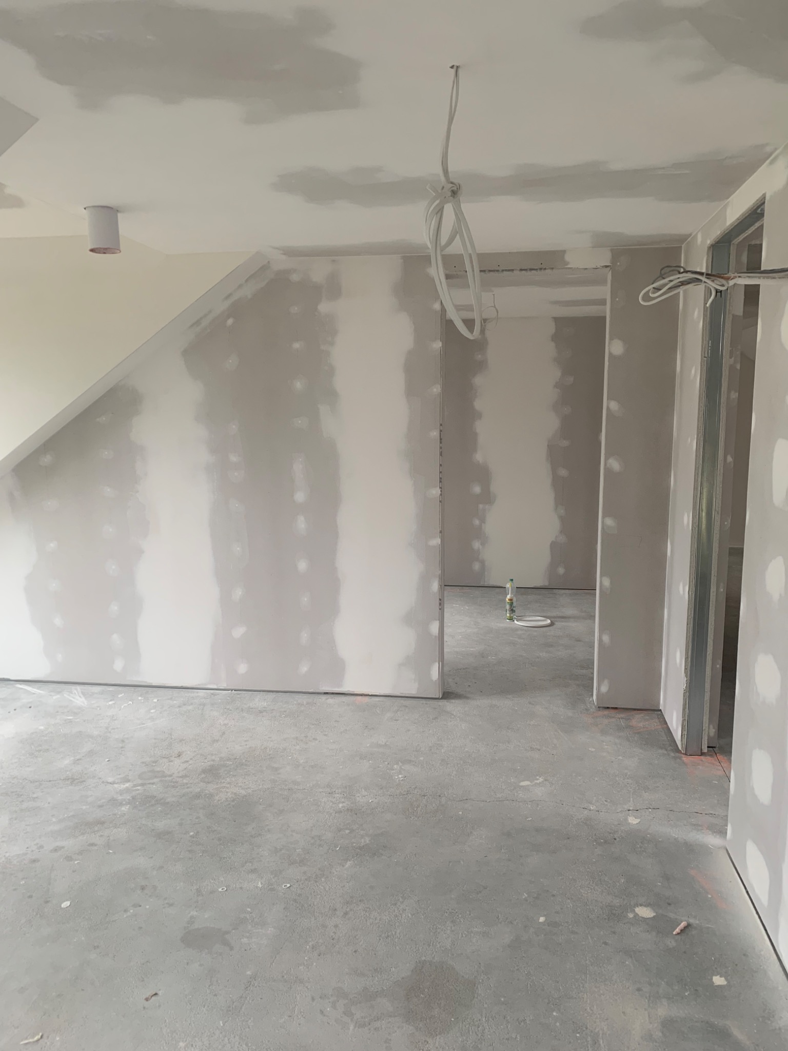 Wnętrze w trakcie remontu: ściany z płyt kartonowo-gipsowych z widocznymi miejscami szpachlowania, surowy beton na podłodze, wiszące kable elektryczne, metalowe ościeżnice drzwiowe.