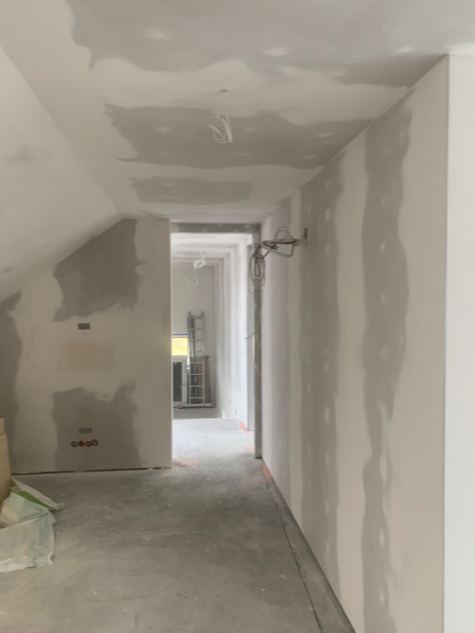 Wnętrze w trakcie prac wykończeniowych: ściany i sufit pokryte warstwą szpachli, widoczne przewody elektryczne, drabina w oddali, podłoga zabezpieczona folią.