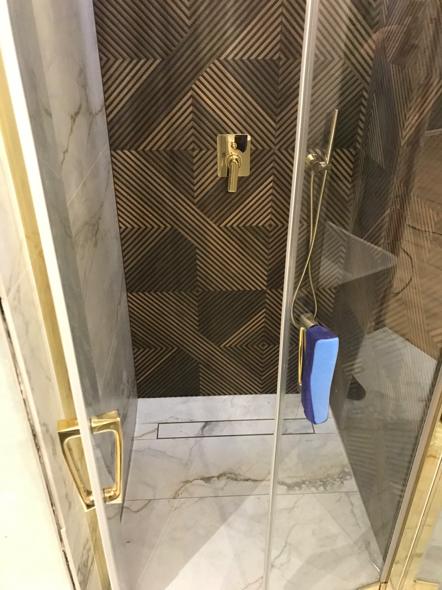 Nowoczesna kabina prysznicowa z marmurową posadzką, złotą armaturą i geometrycznym wzorem na ścianie, widoczna niebieska gąbka na wieszaku.