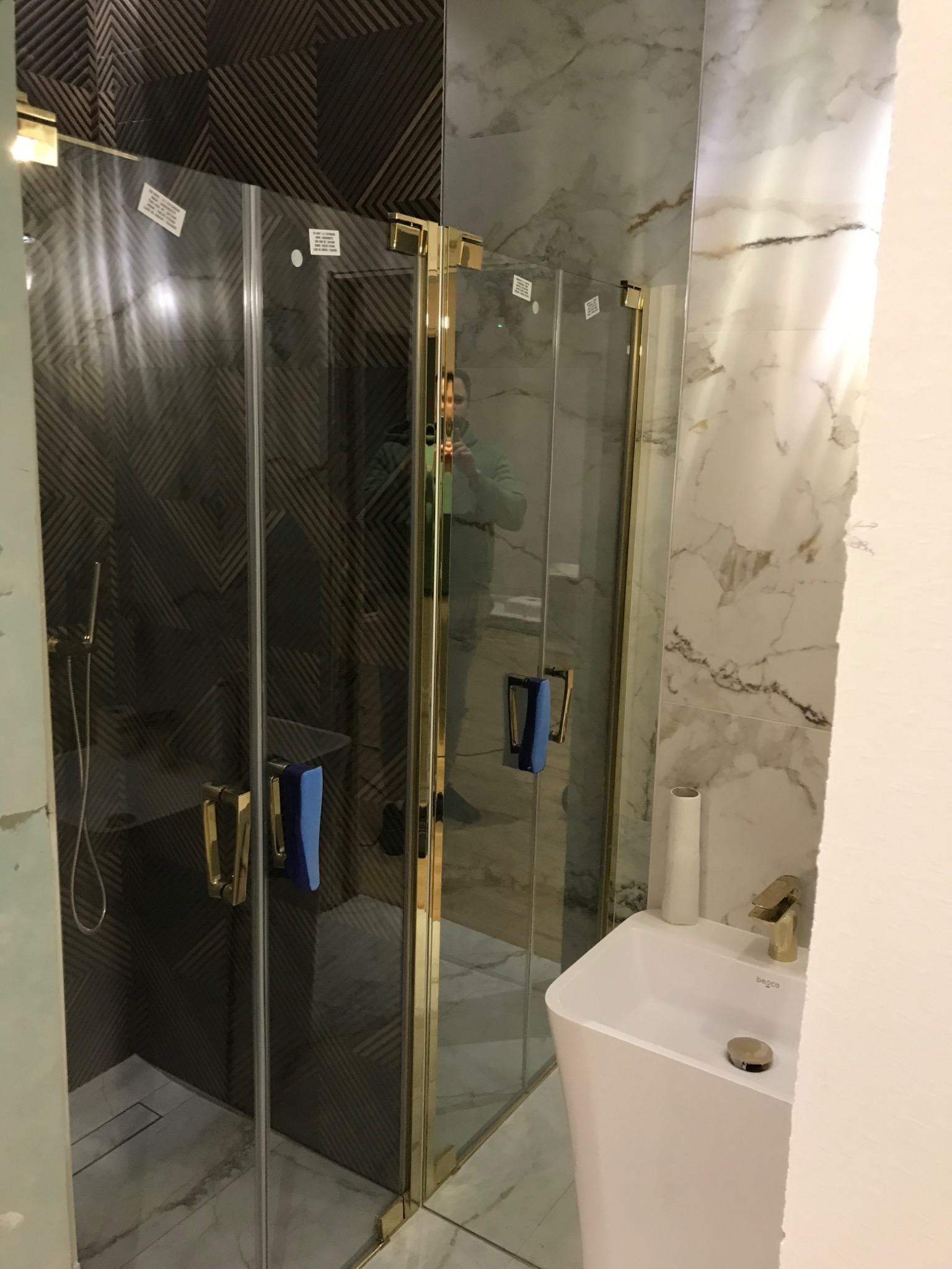 Nowoczesna kabina prysznicowa z złotymi profilami, zamontowana w łazience z marmurowymi i geometrycznymi płytkami, widoczna umywalka wolnostojąca i bateria.