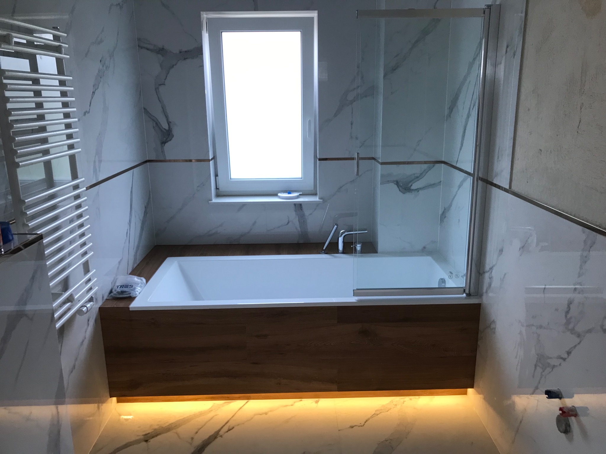 Nowoczesna łazienka z wanną obudowaną drewnem, oświetleniem LED pod wanną, marmurowymi płytkami na ścianach z dekoracyjnymi listwami, szklanym parawanem nawannowym i grzejnikiem drabinkowym.