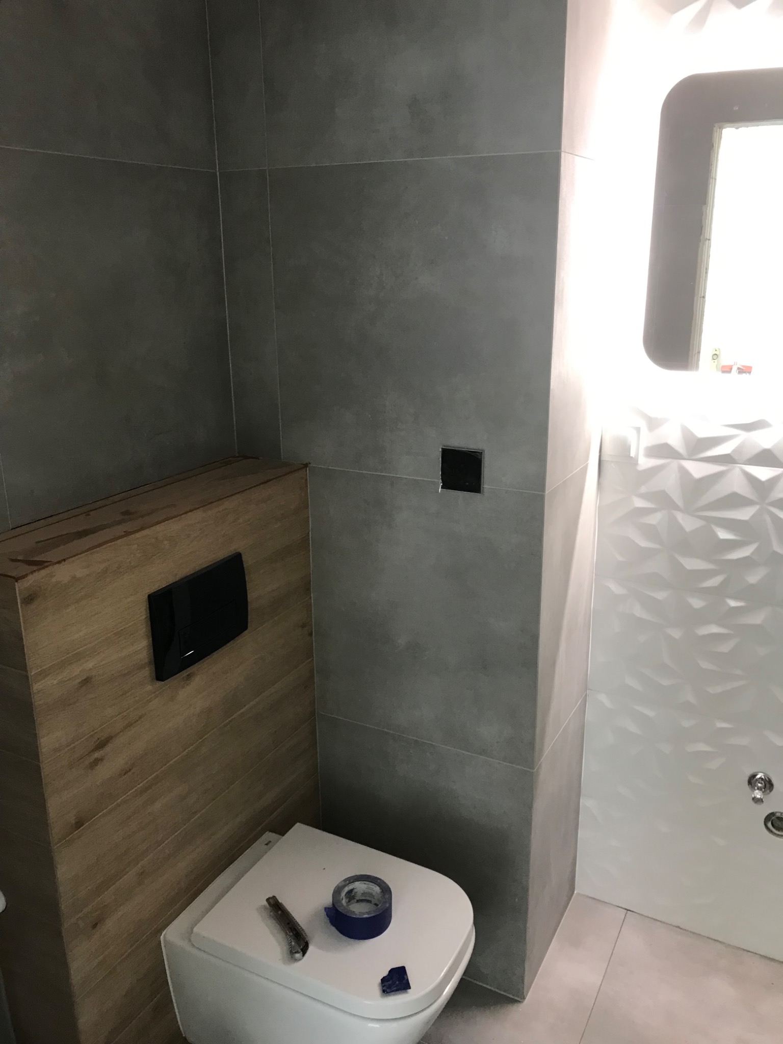 Częściowo wykończona łazienka z szarymi płytkami, drewnopodobną zabudową stelaża WC i białą ścianą z trójwymiarową fakturą. Na desce sedesowej leży nóż tapicerski i rolka niebieskiej taśmy malarskiej.
