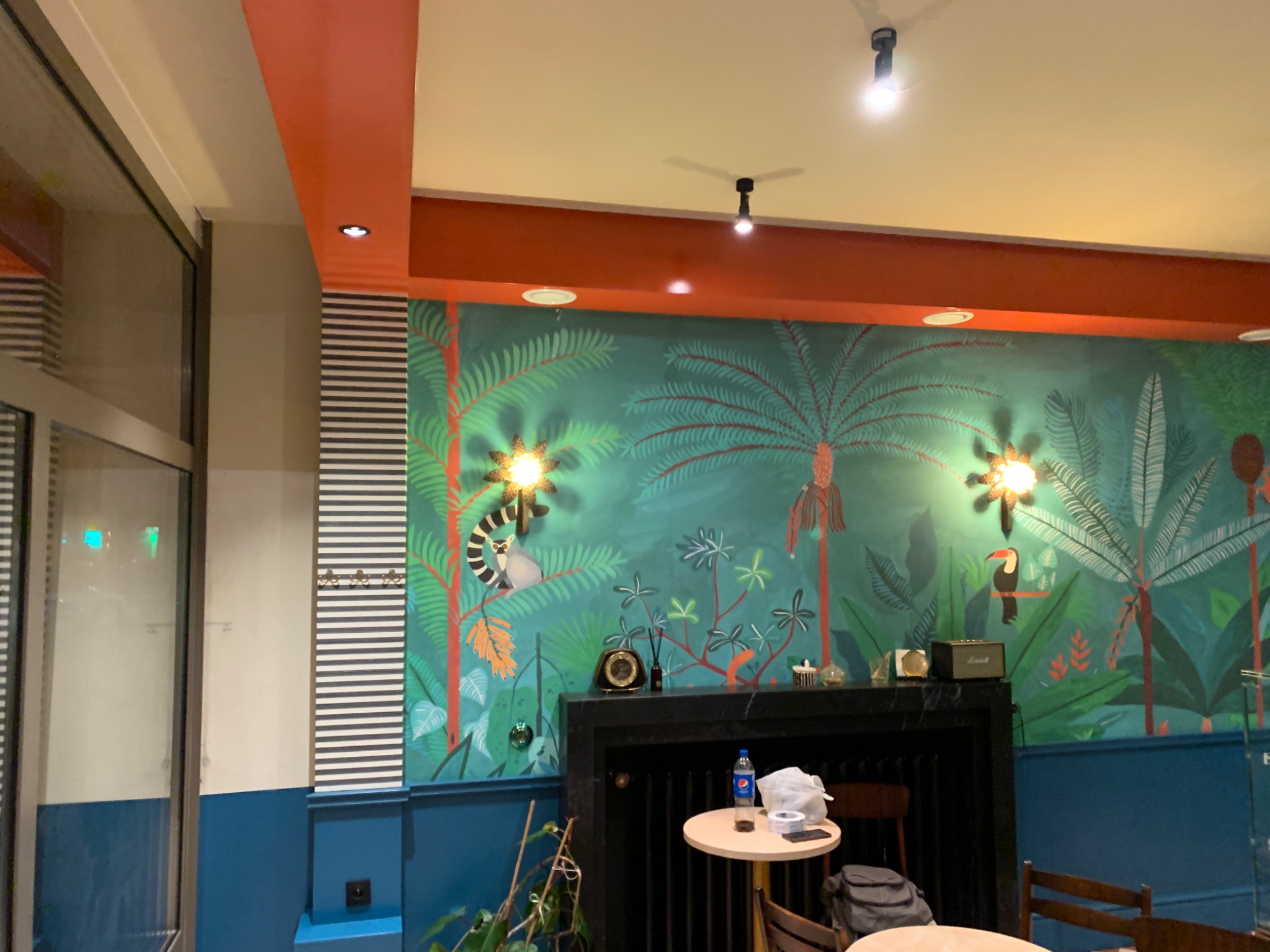 Wnętrze lokalu z dekoracyjną ścianą w tropikalne motywy, pomalowaną na turkusowo z rysunkami palm, tukana i lemura, nad czarnym kominkiem z zegarem i głośnikiem Marshall, oświetlone punktowymi...