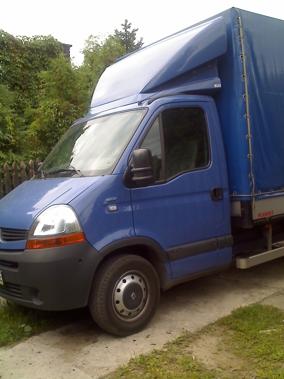 Niebieski samochód dostawczy z plandeką na podwórku, widok z boku na przednią część pojazdu, z widocznym napisem 'PLANDEX' na plandece i zaroślami w tle.