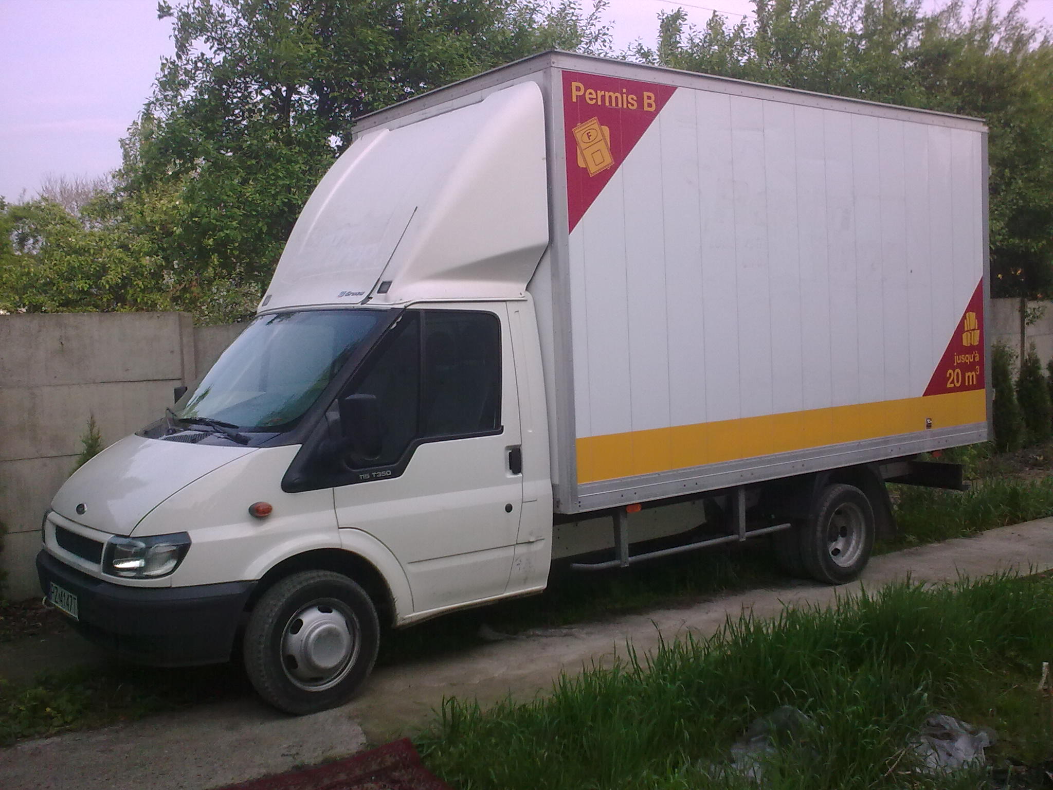 Biały samochód dostawczy Ford Transit z wysoką zabudową, oznaczony informacją o pojemności 20 m3 i dopuszczalnym prawie jazdy kategorii B, zaparkowany na betonowym podłożu w otoczeniu zieleni.