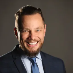 Portret uśmiechniętego mężczyzny w garniturze w kratę i błękitnym krawacie na szarym tle, z wyraźną brodą i starannie ułożonymi włosami.