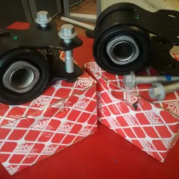 Dwa czarne silentblocki zawieszenia z metalowymi elementami mocującymi, leżące na czerwonych pudełkach marki Febi Bilstein, obok srebrne śruby.