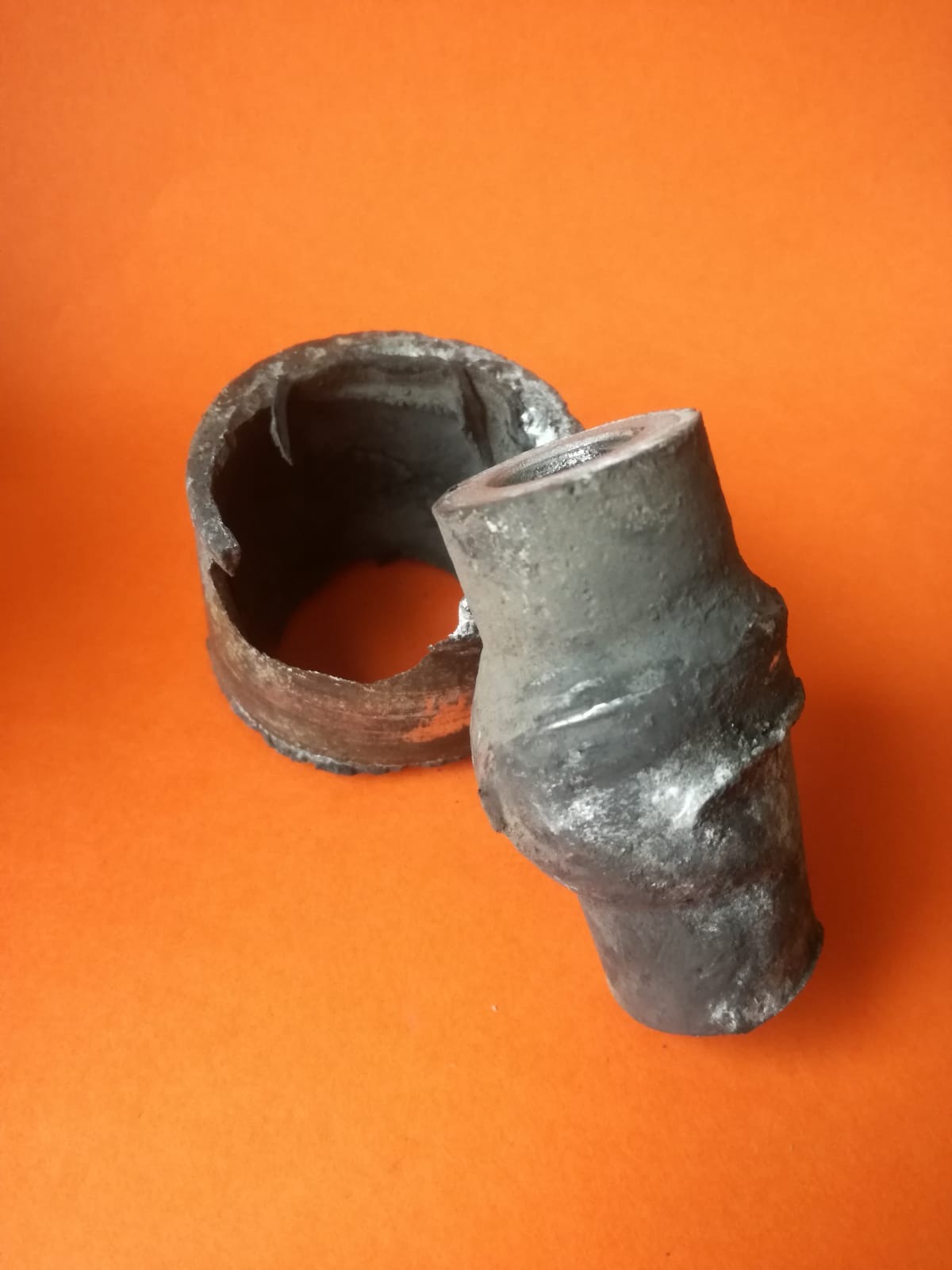Uszkodzony element metalowy, prawdopodobnie rura wydechowa, z widocznymi śladami korozji i pęknięć, na pomarańczowym tle.
