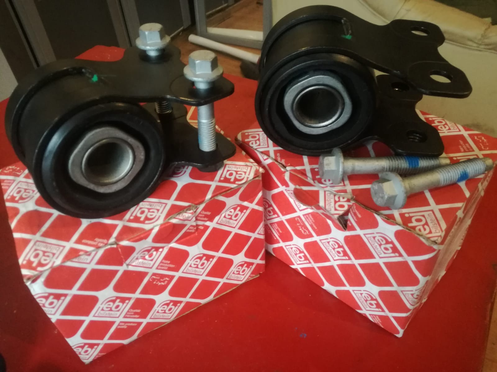 Dwa czarne silentblocki zawieszenia z metalowymi elementami mocującymi, leżące na czerwonych pudełkach marki Febi Bilstein, obok srebrne śruby.