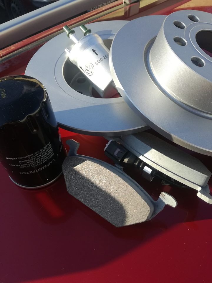 Zestaw nowych części samochodowych na błyszczącym czerwonym tle: tarcze hamulcowe, filtr oleju z napisem LANGZEITFILTER, filtr paliwa marki VW i klocki hamulcowe.