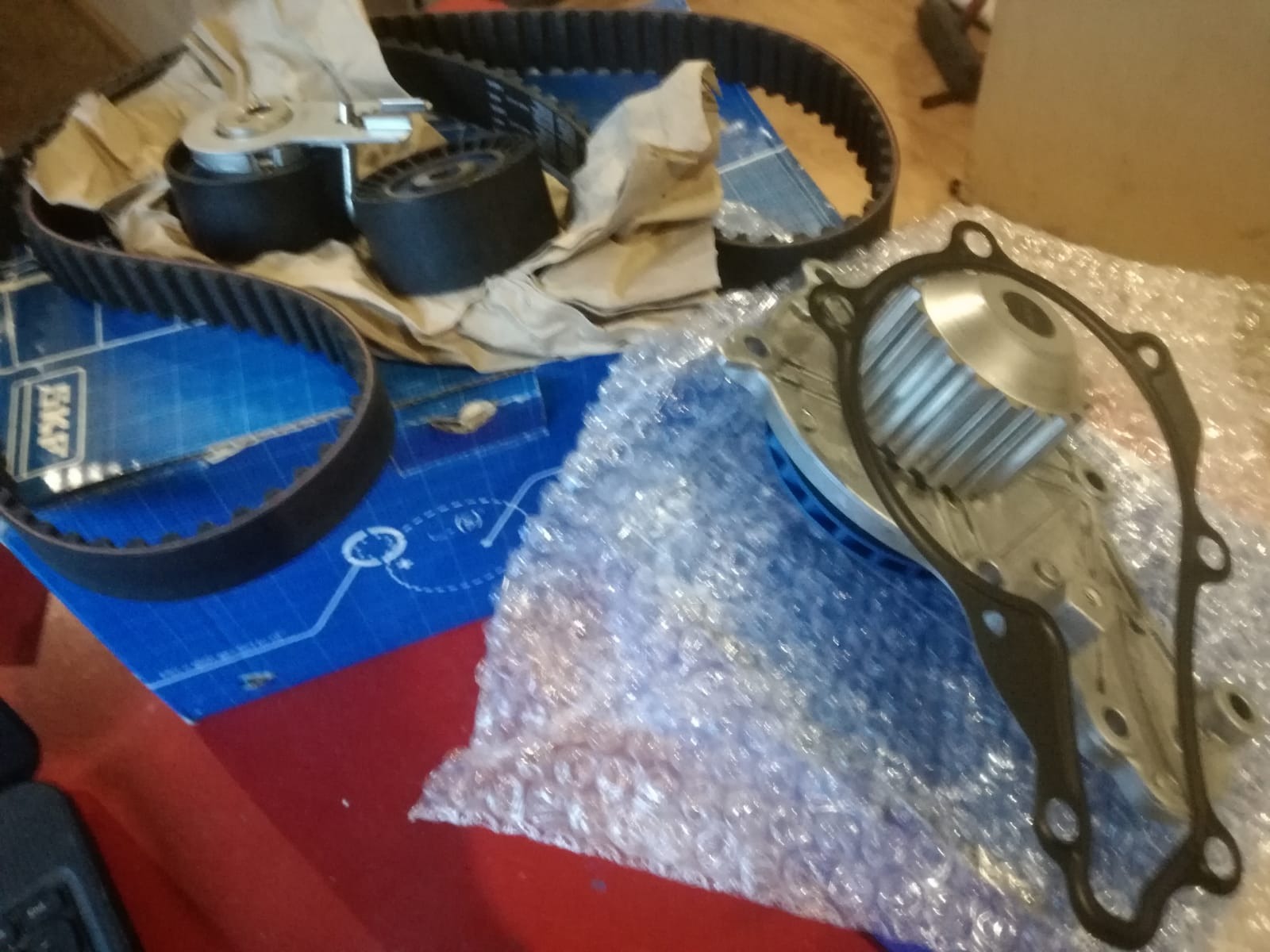 Zestaw części samochodowych: pasek rozrządu, rolki napinające i pompa wody, rozłożone na niebieskim pudełku i folii bąbelkowej przed montażem.