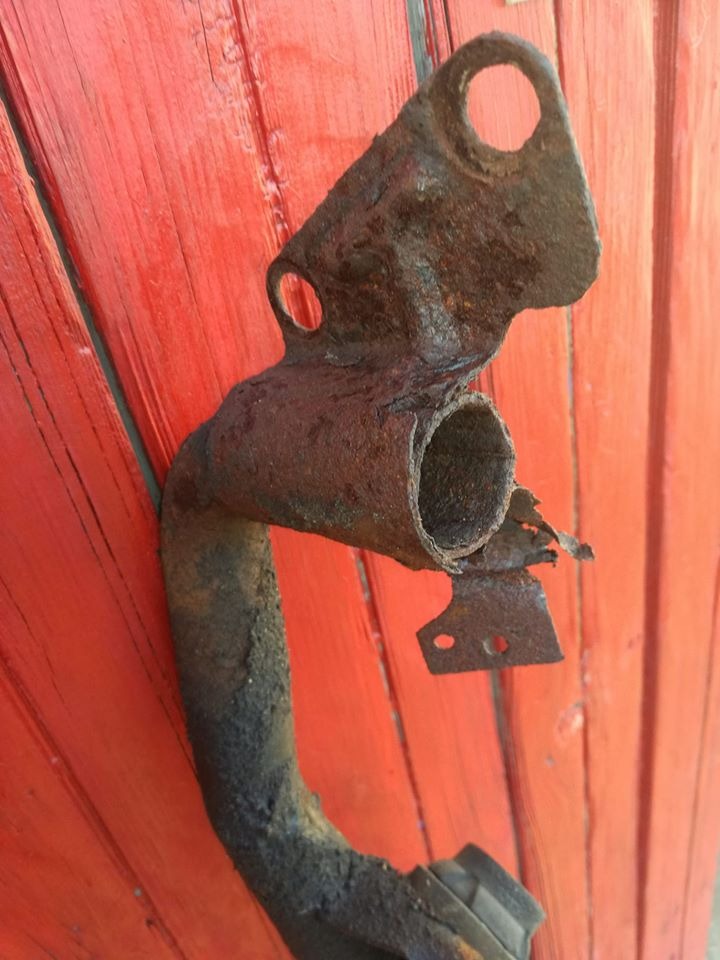 Zardzewiały element układu wydechowego, prawdopodobnie rura, z widocznymi śladami korozji i uszkodzeń, na tle czerwonych desek.