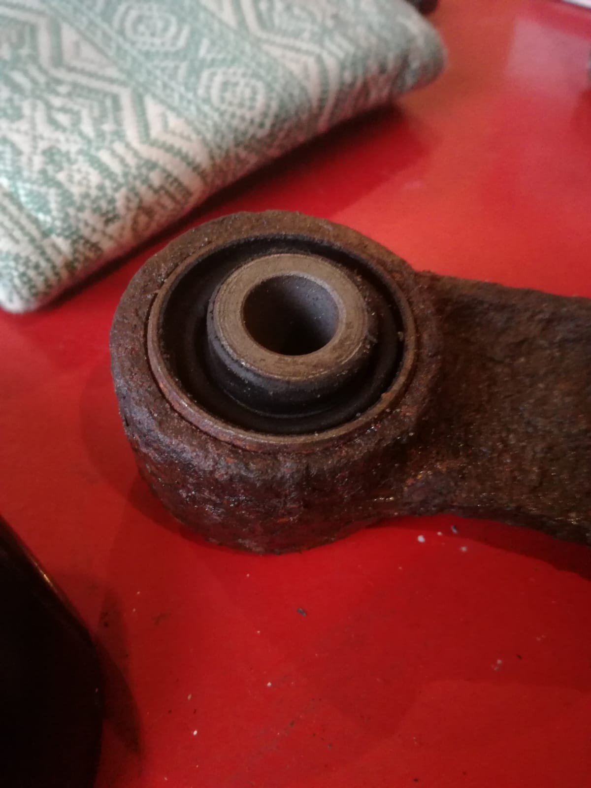 Zardzewiałe metalowe ramię wahacza samochodowego z tuleją gumowo-metalową na czerwonym tle stołu, widoczne ślady korozji.