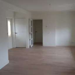 Jasne, przestronne wnętrze z panelami podłogowymi w odcieniach beżu, białe ściany, widoczne drzwi wewnętrzne i fragment zabudowy kuchennej w kolorze grafitowym.