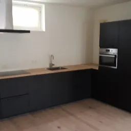 Nowoczesna kuchnia z czarnymi szafkami i drewnianym blatem, widok na zlewozmywak, płytę indukcyjną i piekarnik, oświetlona przez okno