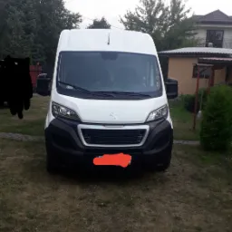 Biały bus Peugeot zaparkowany na trawniku przed domem jednorodzinnym, widok z przodu, zasłonięta tablica rejestracyjna.