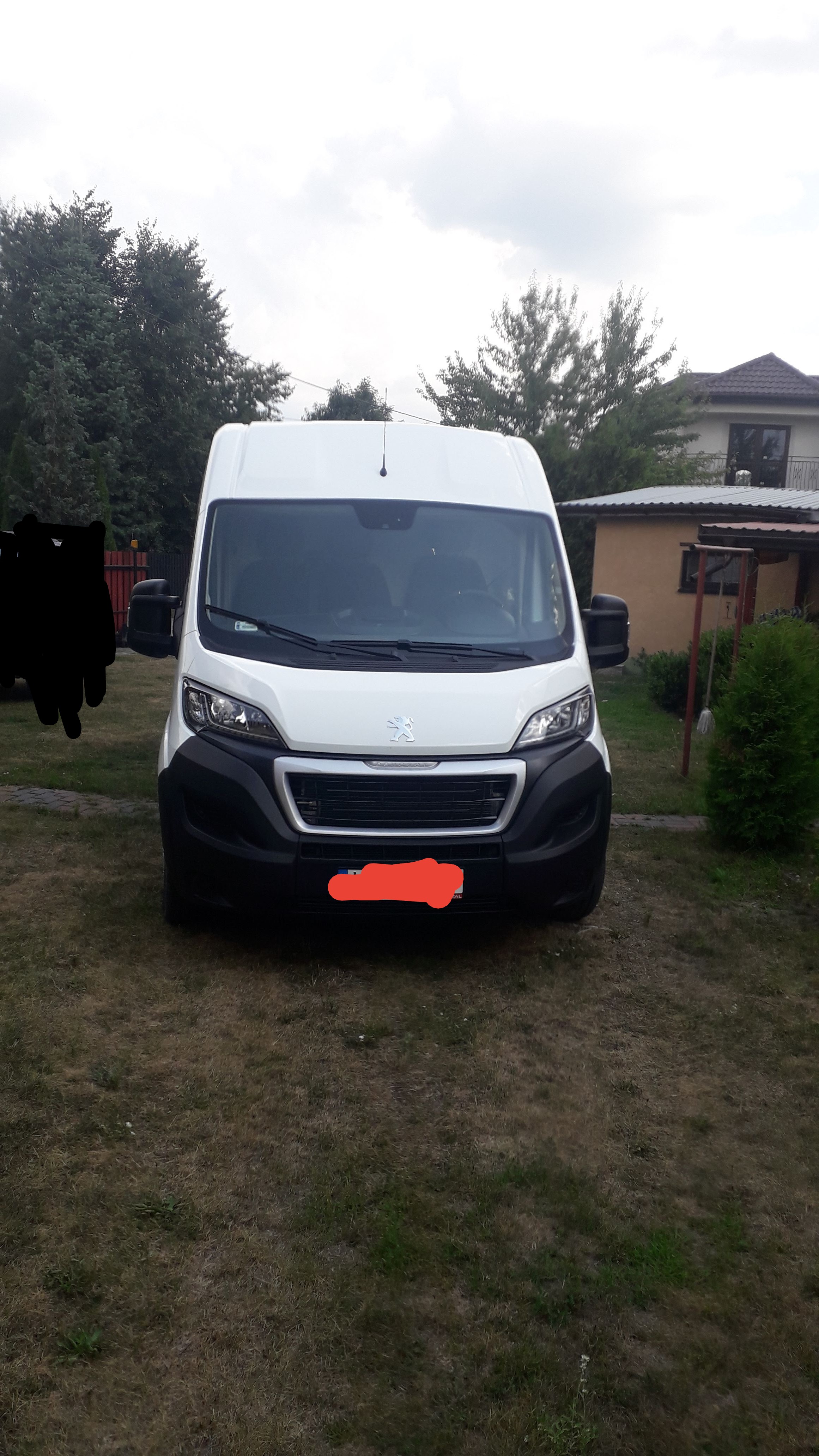 Biały bus Peugeot zaparkowany na trawniku przed domem jednorodzinnym, widok z przodu, zasłonięta tablica rejestracyjna.