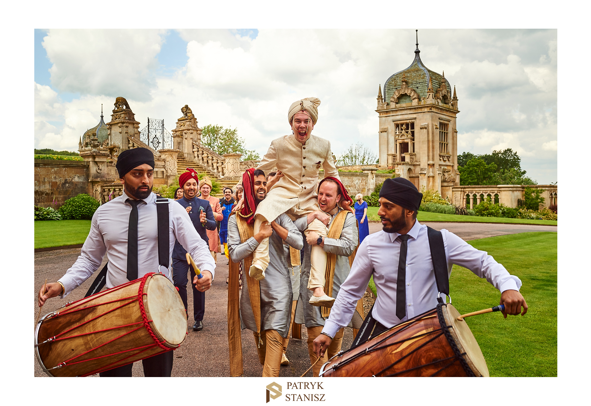 Uśmiechnięty pan młody niesiony na ramionach przez mężczyzn w tradycyjnych strojach, grający na bębnach dhol, w tle zabytkowa budowla i goście weselni.