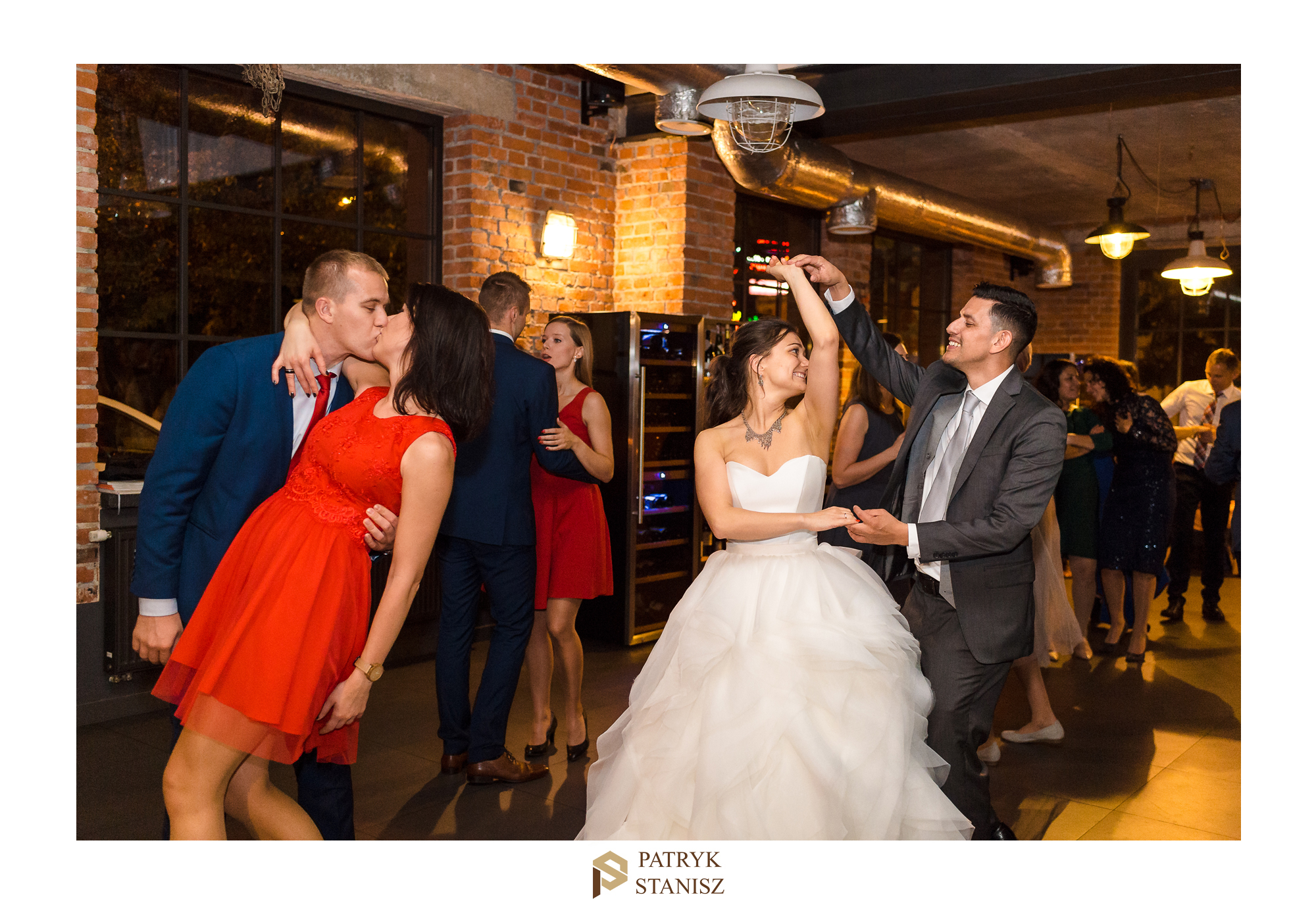 Para młoda tańczy na parkiecie w otoczeniu gości weselnych, ceglana ściana i industrialne lampy w tle.