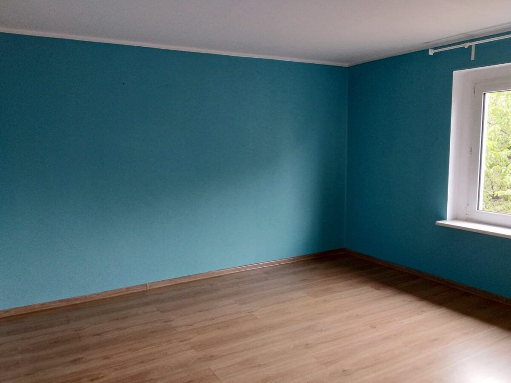 Pomalowane na turkusowo wnętrze pokoju z drewnianą podłogą i białym oknem z widokiem na zieleń.