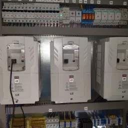 Rozdzielnica elektryczna z zainstalowanymi zabezpieczeniami, stycznikami i falownikami LS H100, oznaczona numerami od 1 do 24, widoczne okablowanie i elementy montażowe.