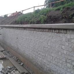 T.M. Europa Plus Sp. z o.o. - Budowa muru oporowego z kostki granitowej, pracownik w kamizelce odblaskowej i kasku kontroluje jakość wykonania, widoczne deski i nierówności terenu.