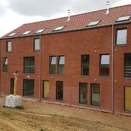 Surowy budynek wielorodzinny z czerwonej cegły z oknami i poddaszem, widok z placu budowy z widocznymi śladami opon i stertą kostki brukowej w tle.