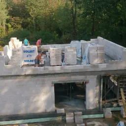 Wznoszenie ścian parterowego domu z białych bloczków Solbet, widoczni pracownicy w trakcie murowania, w tle zalesiony teren.