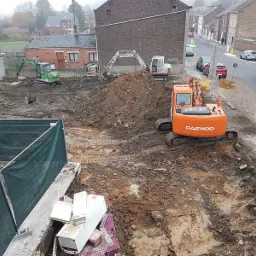 Plac budowy z kilkoma koparkami, w tym pomarańczową Daewoo, przygotowujący teren pod budowę w zabudowie miejskiej, widok z góry.