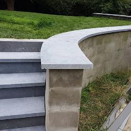 T.M. Europa Plus Sp. z o.o. - Schody zewnętrzne z szarej kostki brukowej prowadzące do podwyższonego trawnika z zakrzywionym murem oporowym wykończonym szarym kamieniem; w tle zaparkowany biały samochód dostawczy.