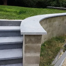 Schody zewnętrzne z szarej kostki brukowej prowadzące do podwyższonego trawnika z zakrzywionym murem oporowym wykończonym szarym kamieniem; w tle zaparkowany biały samochód dostawczy.