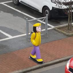 T.M. Europa Plus Sp. z o.o. - Kolorowa, plastikowa figura pieszego w żółtej kurtce, fioletowych spodniach i czapce, stojąca na chodniku z kostki brukowej przy ulicy i parkingu.
