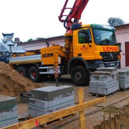 T.M. Europa Plus Sp. z o.o. - Żółta betonomieszarka z pompą LEMAR na placu budowy, obok sterty kostki brukowej ułożonej na paletach, w tle biało-zielona betoniarka i budynek z czerwonymi drzwiami.