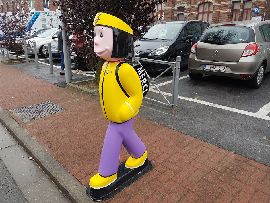 Kolorowa, plastikowa figura chłopca w żółtej kurtce i fioletowych spodniach z napisem 'MERCI' na plecach, stojąca na chodniku z kostki brukowej na tle parkingu i budynku.