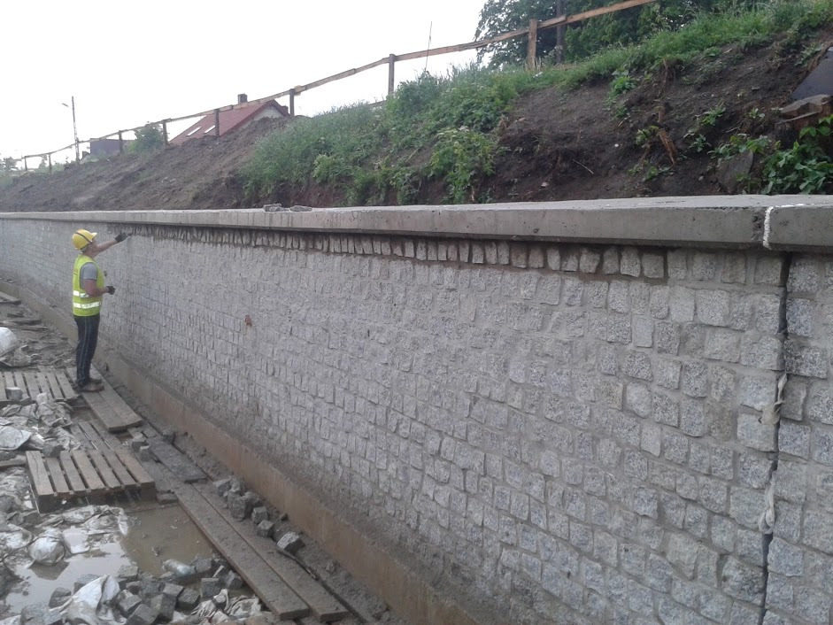 Budowa muru oporowego z kostki granitowej, pracownik w kamizelce odblaskowej i kasku kontroluje jakość wykonania, widoczne deski i nierówności terenu.