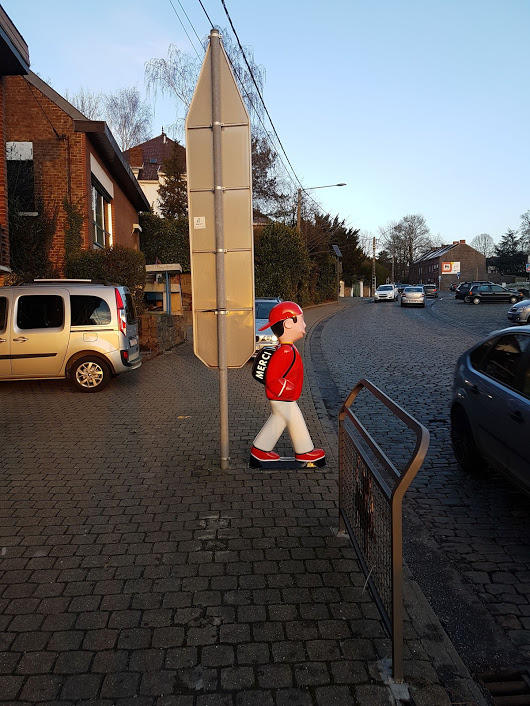 Manekin pieszego w czerwonym stroju i czapce, stojący na chodniku z kostki brukowej obok znaku drogowego, w tle zabudowa jednorodzinna i zaparkowane samochody.