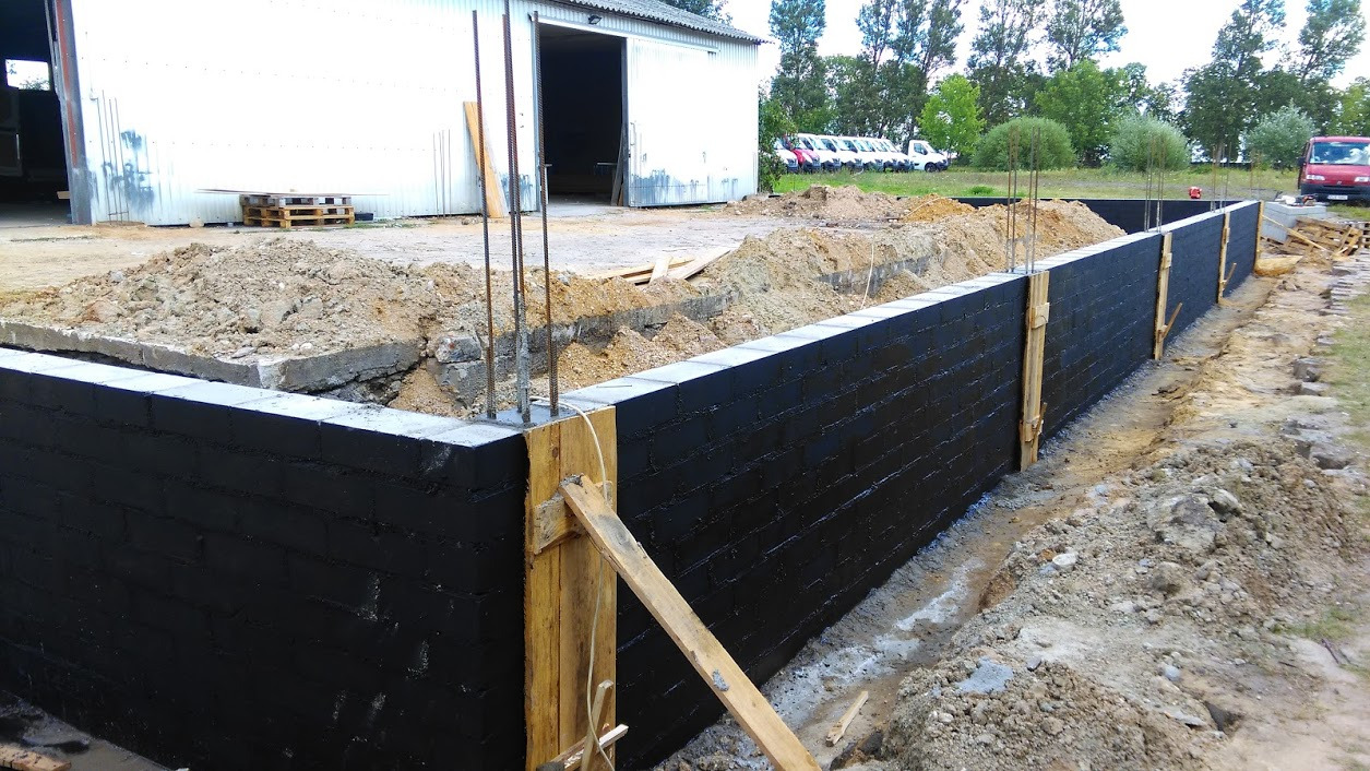 Wznoszenie fundamentów hali przemysłowej z bloczków betonowych, zabezpieczonych czarną hydroizolacją, widoczne szalunki i wystające pręty zbrojeniowe, w tle budynek magazynowy z blachy trapezowej...
