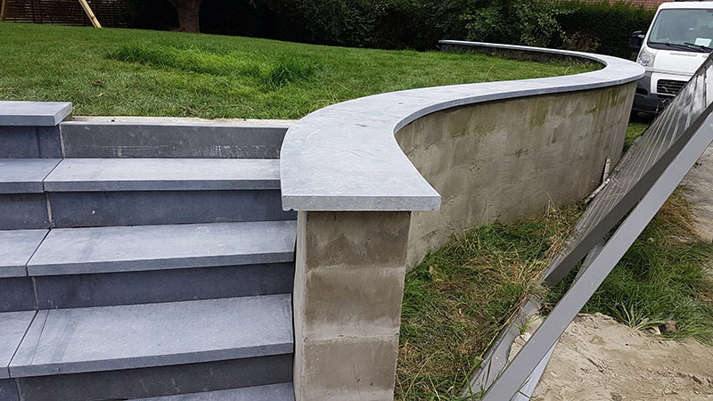 Schody zewnętrzne z szarej kostki brukowej prowadzące do podwyższonego trawnika z zakrzywionym murem oporowym wykończonym szarym kamieniem; w tle zaparkowany biały samochód dostawczy.