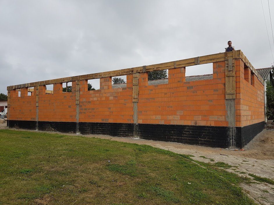 Wznoszona ściana hali z czerwonej cegły, z widoczną hydroizolacją fundamentów i drewnianymi elementami konstrukcyjnymi, w tle pochmurne niebo i sylwetka mężczyzny na szczycie ściany.