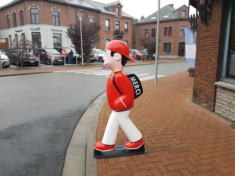 Figurka chłopca w czerwonej czapce i kurtce z napisem 'MERCI' na plecaku, ustawiona na chodniku z kostki brukowej w otoczeniu miejskiej zabudowy.