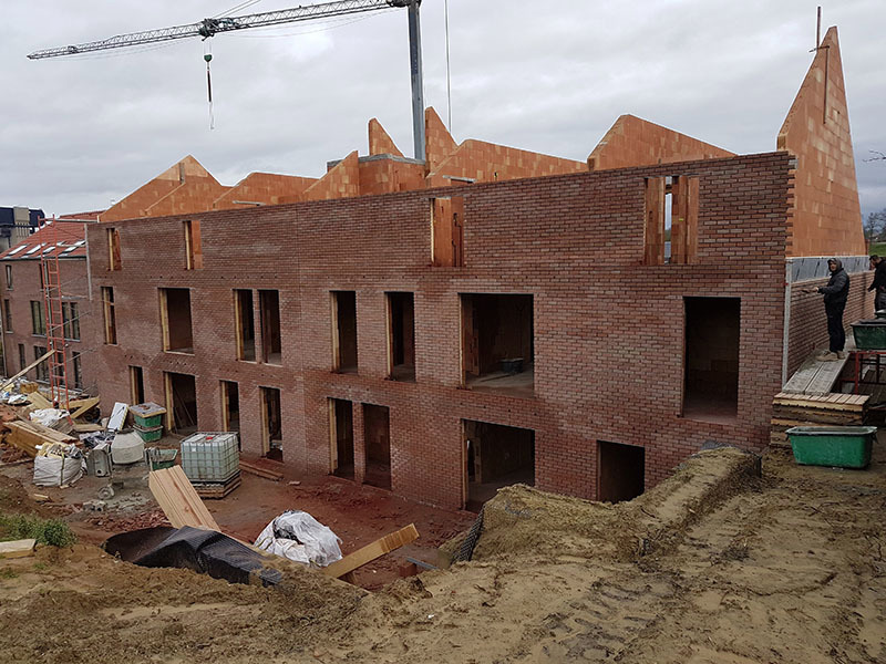 Wznoszenie dwukondygnacyjnego budynku mieszkalnego z czerwonej cegły, widoczny żuraw budowlany i dwóch pracowników przy murowaniu ściany szczytowej.