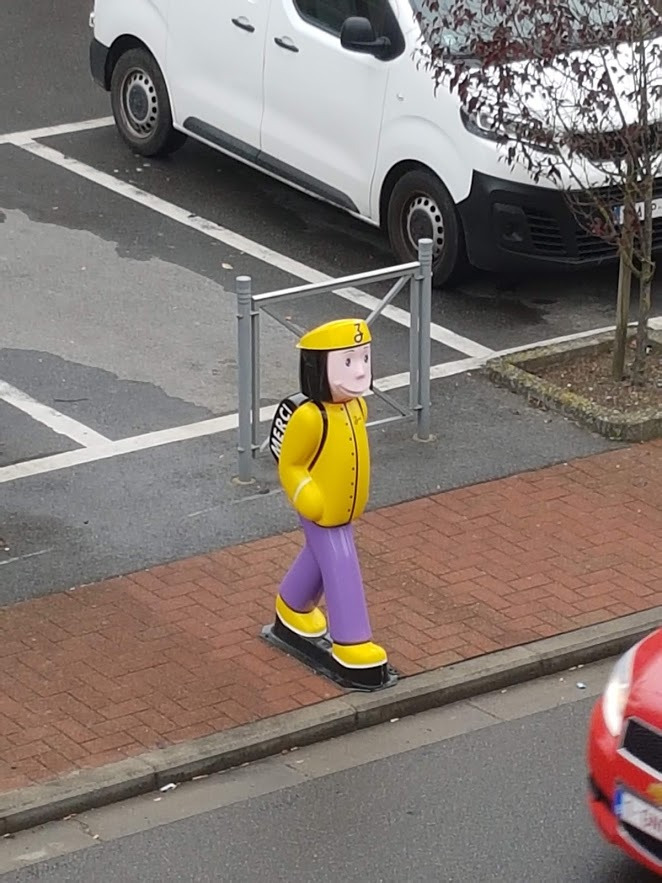 Kolorowa, plastikowa figura pieszego w żółtej kurtce, fioletowych spodniach i czapce, stojąca na chodniku z kostki brukowej przy ulicy i parkingu.